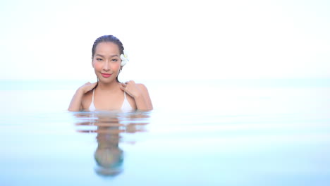 Close-up,-rule-of-thirds,-push-in-of-a-beautiful-young-woman-with-flawless-skin-and-a-flower-behind-her-left-ear-standing-chest-high-in-still-pool-water