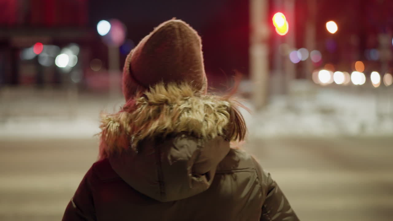 First-person view of a woman in a winter coat with a fur hood, holding her head with both hands, standing at a nighttime urban intersection with blurry lights and red traffic signal in the background