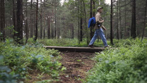 Young-man-with-camera-walks-through-forest,-pan-from-behind-bushes