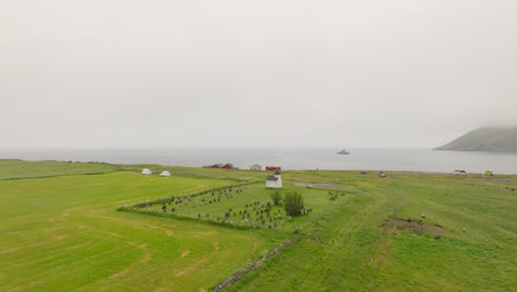 Aerial-View-Of-Birds-Flying-Over-Unstad-Chapel-And-Unstad-Cemetery-On-A-Foggy-Day-In-Vestvagoya,-Norway