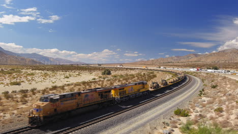 Vista-Aérea-Del-Tren-Que-Transporta-Cientos-De-Vehículos-Militares-Estadounidenses,-Tanques-Y-Humvees-Son-Transportados-Por-Ferrocarril-A-Través-Del-Desierto-Fuera-De-Palm-Springs