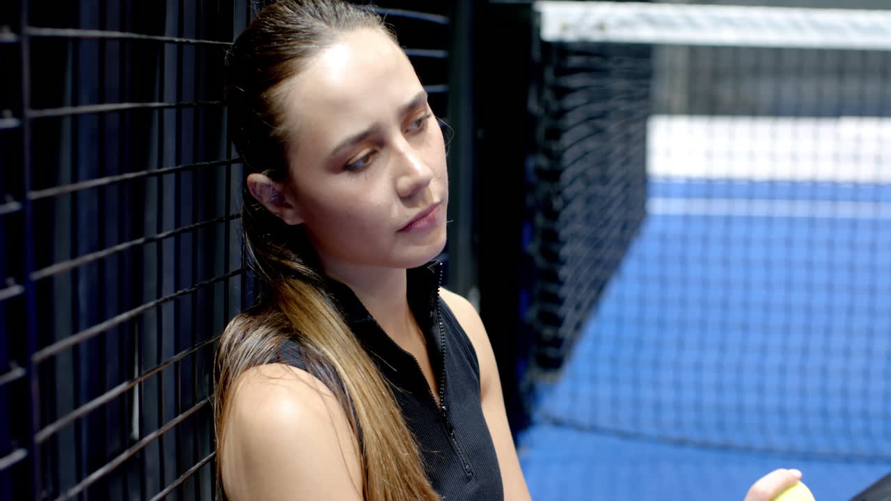Focused woman resting against padel on indoor court fence, preparing for next match