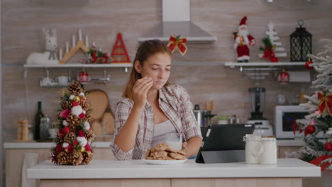 Portrait-of-children-eating-baked-delicious-cookies-drinking-milk-while-watching-online-xmas-video-on-tablet