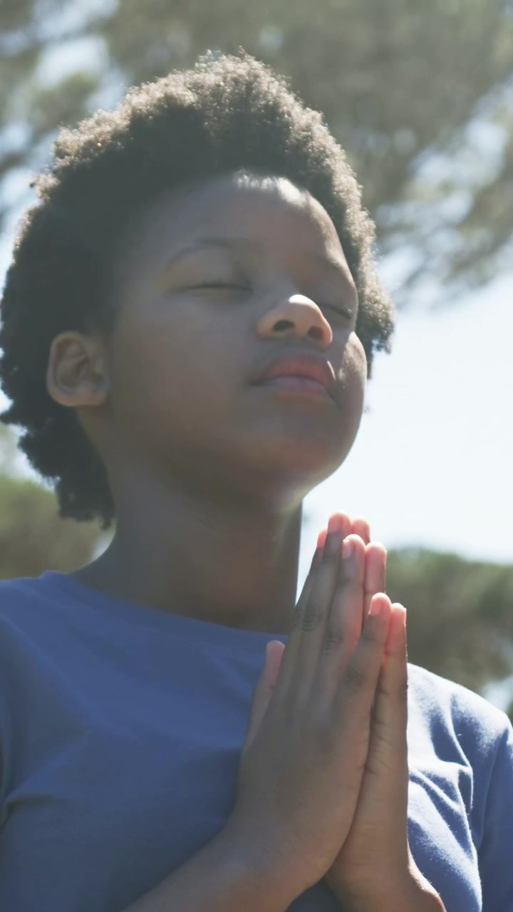 Vertical video: Praying African youth hands pressed as camera pulling back in sunny park blue shirt