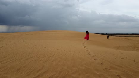 Raven-hair-woman-in-long-red-dress-runs-with-dog-on-remote-sand-dune