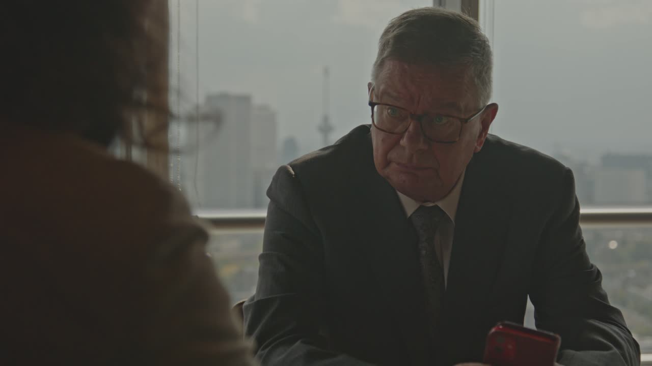 An older businessman sits across a business partner in an office and talks
