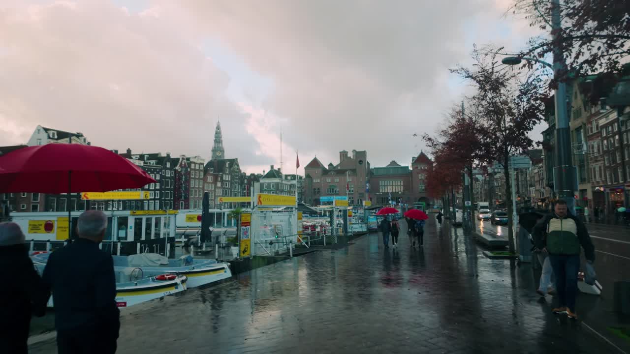 Rainy scene at Amsterdam's Damrak featuring boats docked by the canal, pedestrians walking with umbrellas, and historic buildings under a cloudy sky. Location: Amsterdam, Netherlands