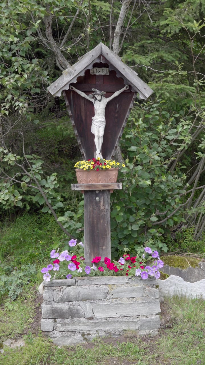Una croce cristiana di legno su una base circondata da fiori, cespugli ed erba all'aperto.
