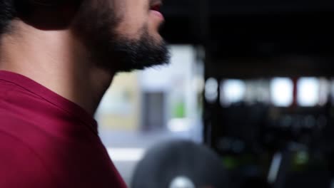 Joven-Hombre-Barbudo-Caucásico-Con-Sombrero-Y-Auriculares-Haciendo-Ejercicio-De-Bíceps-Con-Pesas-En-El-Gimnasio,-Perfil-De-Cierre