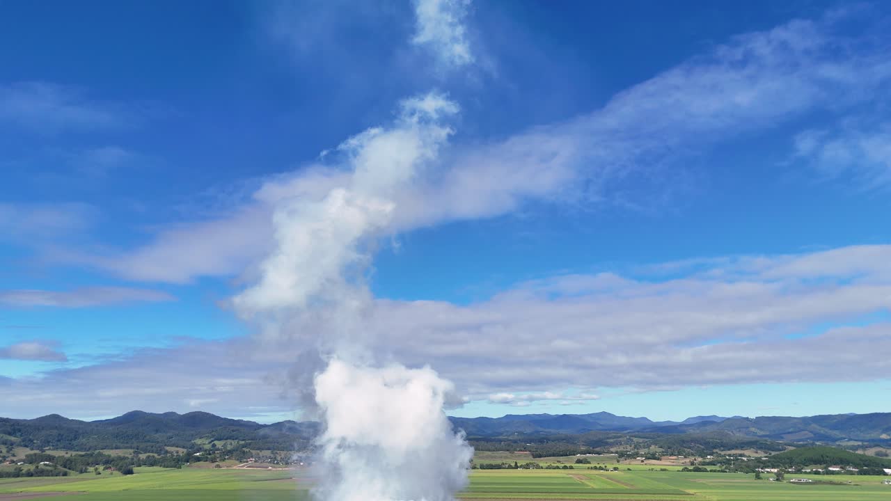 Drone captures steam rising from a smoke stack against a clear blue sky over expansive farmland