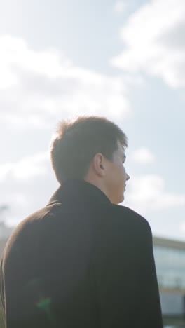 검은 코트를 입은 남자가 은 날 자신감 있게 고, 빛이 반사되고, 도시 배경에 유리 건물이 있으며, 전문성과 현대적인 비즈니스 라이프 스타일을 상징합니다.