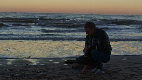 Kaukasisches-Männliches-Mannmodell-Sitzt-In-Der-Hocke-Und-Greift-Mit-Der-Hand-In-Den-Sand-Am-Strand-In-Der-Nähe-Des-Meeresozeans-Am-Sandufer-Mit-Sonnenuntergang-Und-Filmischem-Schauspielerspiel