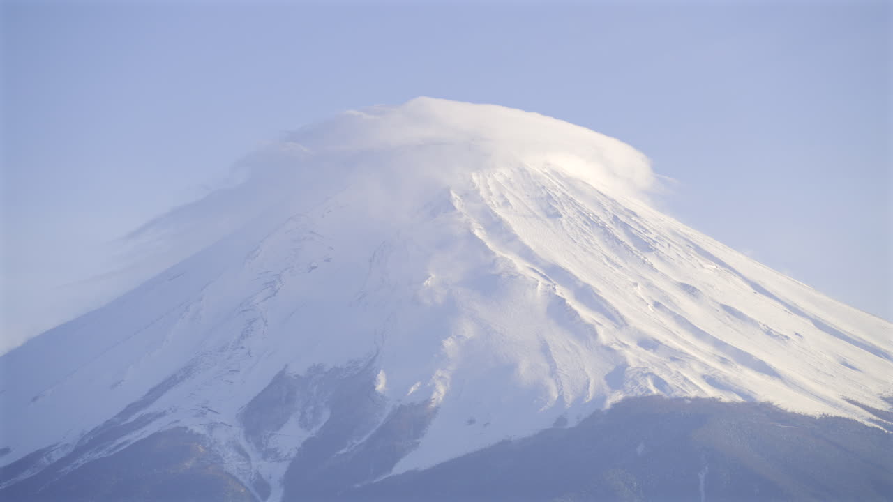A breathtaking sight of a snow-covered volcano as clouds gracefully swirl around its peak during the sunset light, showcasing nature's beauty in serene surroundings