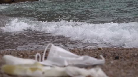 Gebrauchte-Maske-Und-Handschuh-Am-Strand-Mit-Waschwelle-Dahinter,-Herauszoomen-Fokussieren-Hintergrund