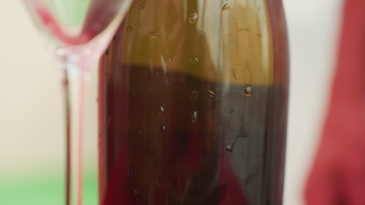 Primo piano di una bottiglia di vino con bicchiere, vino rosso fresco che aderisce alla bottiglia marrone con goccioline di condensa, stelo sottile del calice riflesso a sinistra, tessuto rosso leggermente sfocato e sfondo verde del tavolo