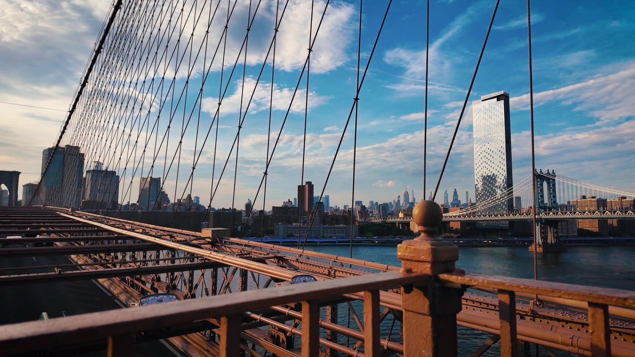 Vista al rallentatore dal Ponte di Brooklyn con il Ponte di Manhattan, lo skyline di Lower Manhattan e l'East River durante l'ora d'oro con nuvole drammatiche e una calda luce serale