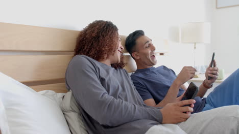 Happy-diverse-gay-male-couple-sitting-on-bed-using-smartphones,-talking-in-the-morning,-slow-motion