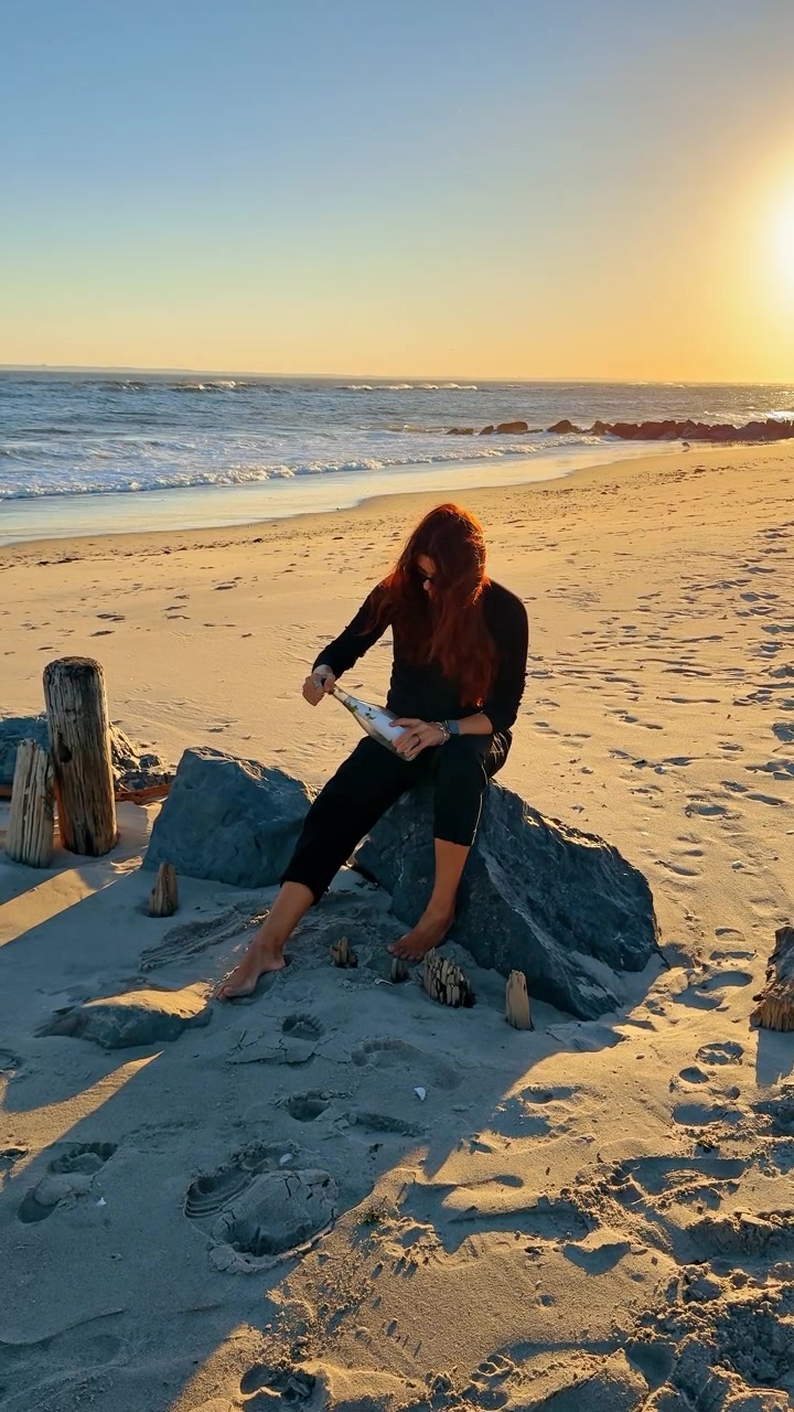 Donna che apre un messaggio in bottiglia. Una donna siede sulla spiaggia aprendo un messaggio in bottiglia durante l'ora d'oro, creando un'atmosfera calma e riflessiva.