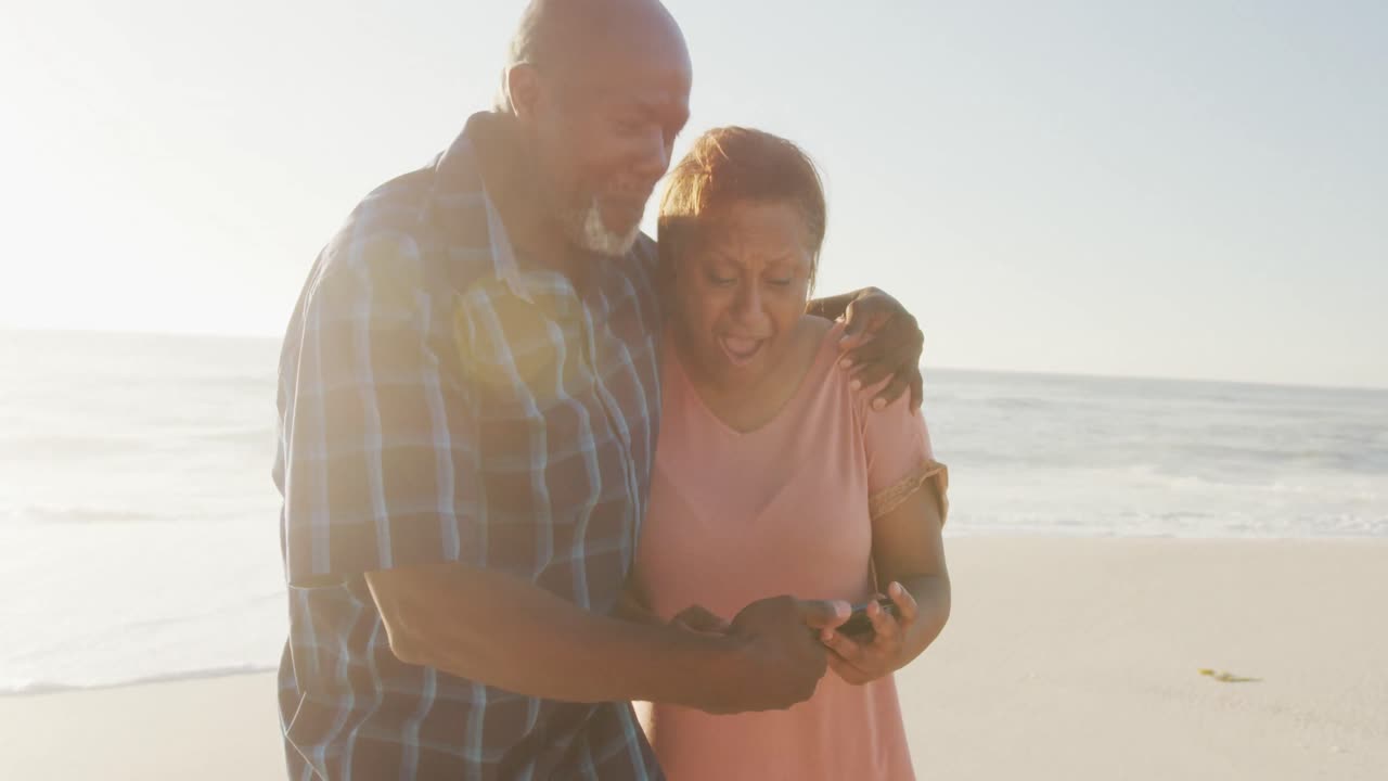 una pareja de ancianos afroamericanos tomados de la mano y usando un teléfono inteligente en una playa soleada