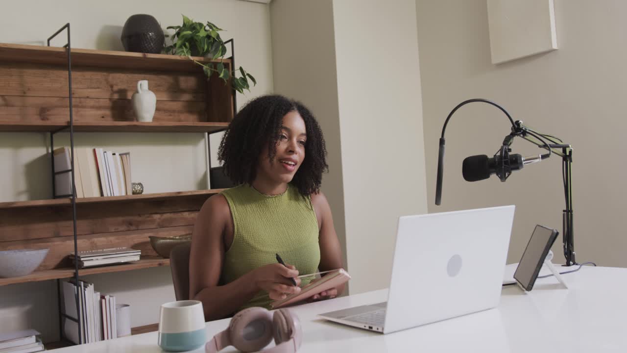 African American woman at home answering offscreen host typing noting in notebook with pen by mic