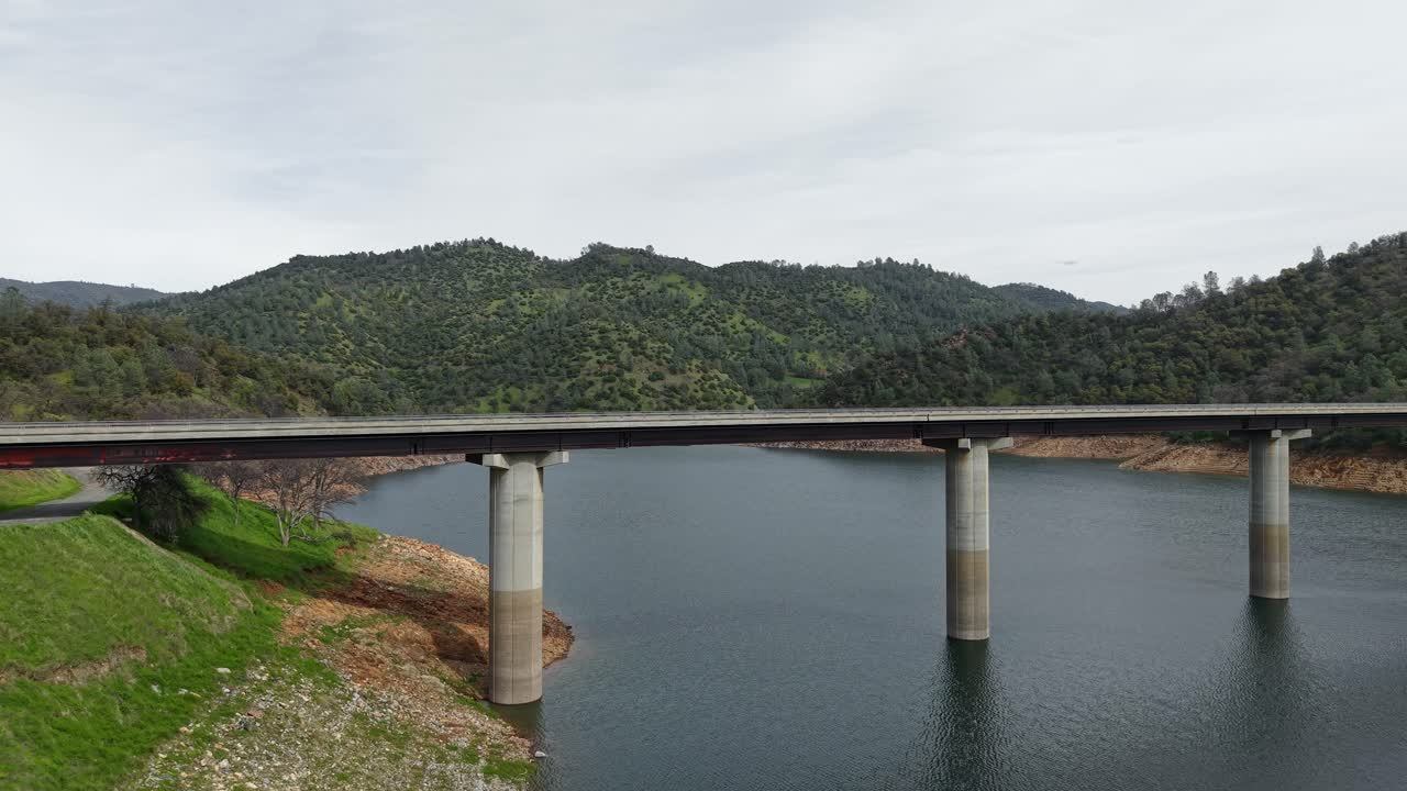 Aerial drone footage captures the Don Pedro Reservoir Bridge in Jamestown, California, stretching across the water with a scenic backdrop of rolling hills and a peaceful, reflective lake surface.