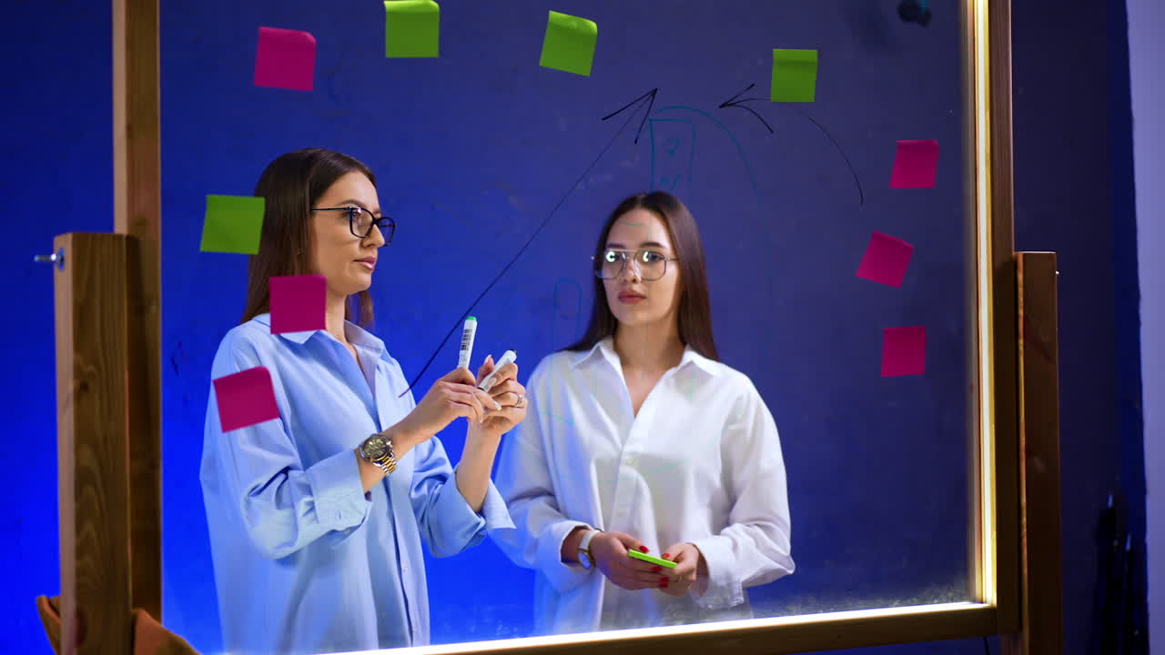 Busy ladies are standing at the glass board in the office. Colleagues draw graphs and put stickers on the glass. Ideas and plans searching.