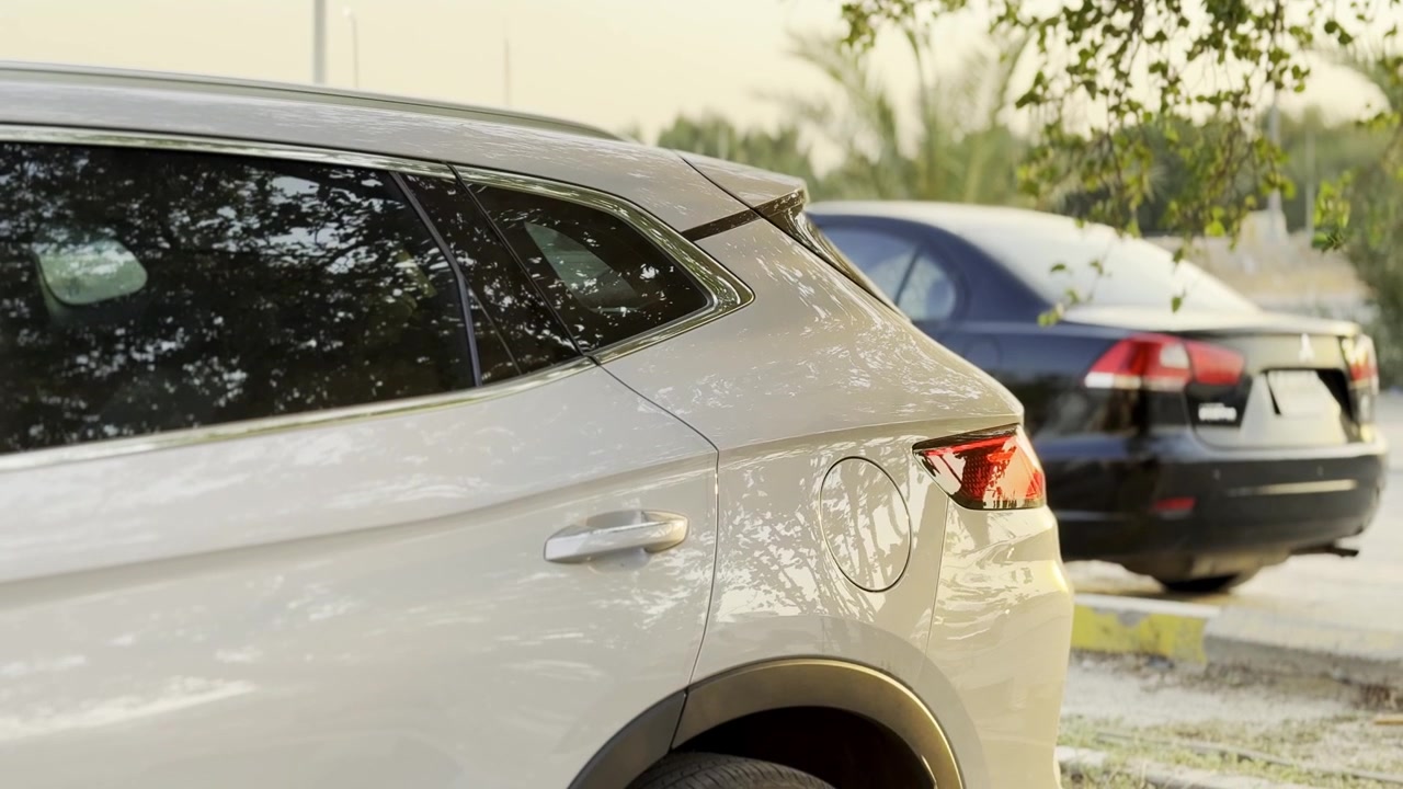Un primo piano di un SUV argentato parcheggiato in un piazzale all'aperto durante l'ora d'oro. I finestrini oscurati del veicolo riflettono gli alberi vicini, mentre un'auto nera rimane sfocata sullo sfondo