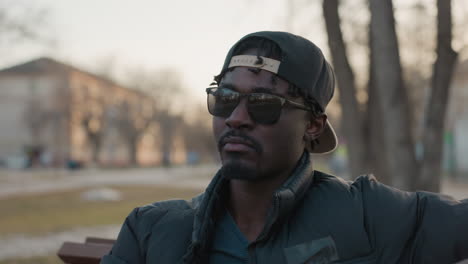 young man in sunglasses sits on park bench sipping juice from bottle, cap worn backward, relaxed posture, sunset glow, blurred people and trees behind