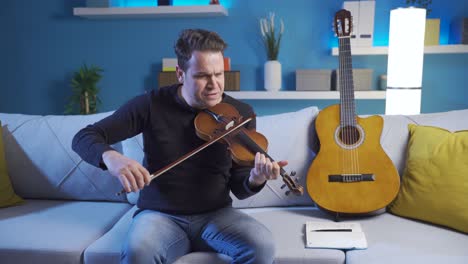 male musician sitting on sofa at home, playing violin and guitar and writing lyrics in notebook while composing new song.