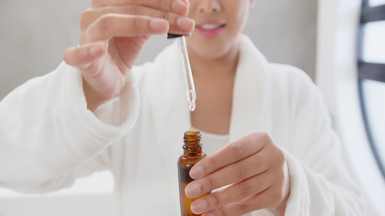 Using dropper, asian woman in white robe applying serum from amber bottle, at home
