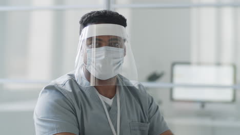 Male-Doctor-in-Face-Shield-and-Mask-Posing-for-Camera-at-Work