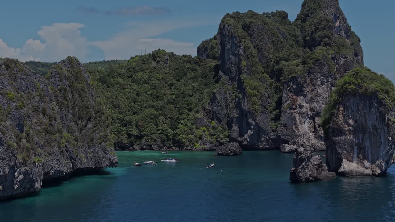 Aerial drone zoom-in of boats gliding near lush coastal cliffs in Phi Phi Islands, Thailand. Shot during bright midday conditions in April.
