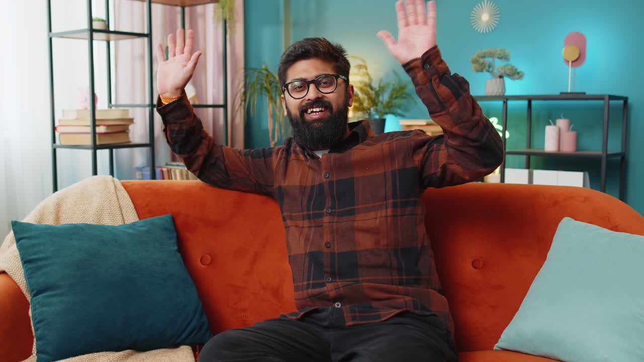 hombre indio sonriendo amistosamente a la cámara agitando las manos gestando hola hola saludando en casa en el sofá