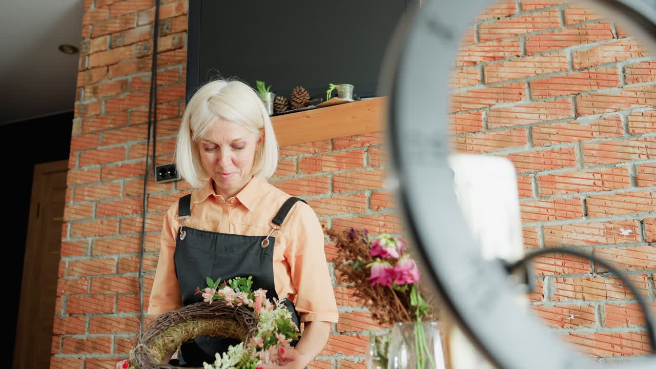 Female florist in black apron holding handmade floral wreath decorated with greenery and blossoms, working in cozy studio with rustic brick wall, dried flowers and plants creating natural decoration