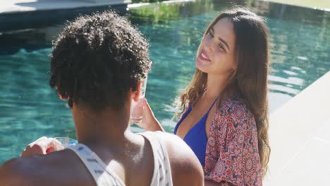 happy biracial couple talking with drinks at pool in garden on sunny day