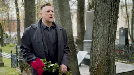 man in black raincoat and suit holding roses while walking in a graveyard 1