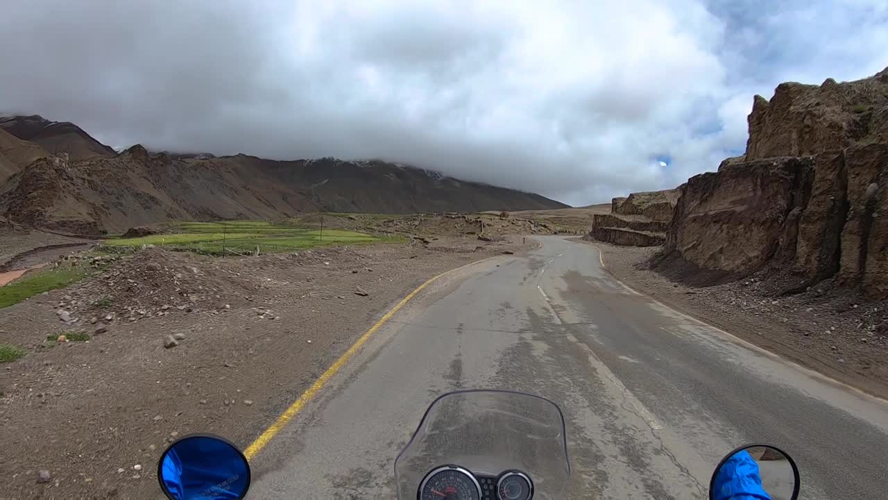 A bike slow riding on a mountain road toward from a barren region toward a distant green Himalayan village. Shot from biker’s point of view.