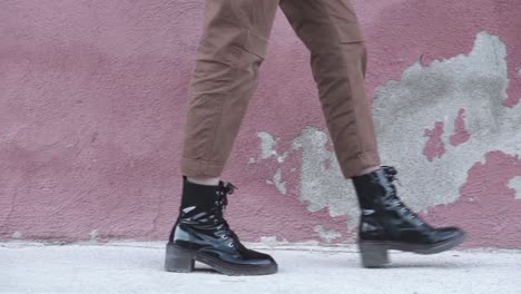 slow motion close up of a woman walking forward wearing shiny black boots and brown pants at an urban set up with a red wall as a background