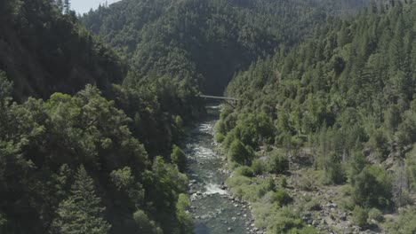 Slow-forward-drone-flight-over-a-rocky-river-toward-a-tall-bridge