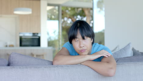 thoughtful teenage asian boy rests his chin on his arms atop a couch at home