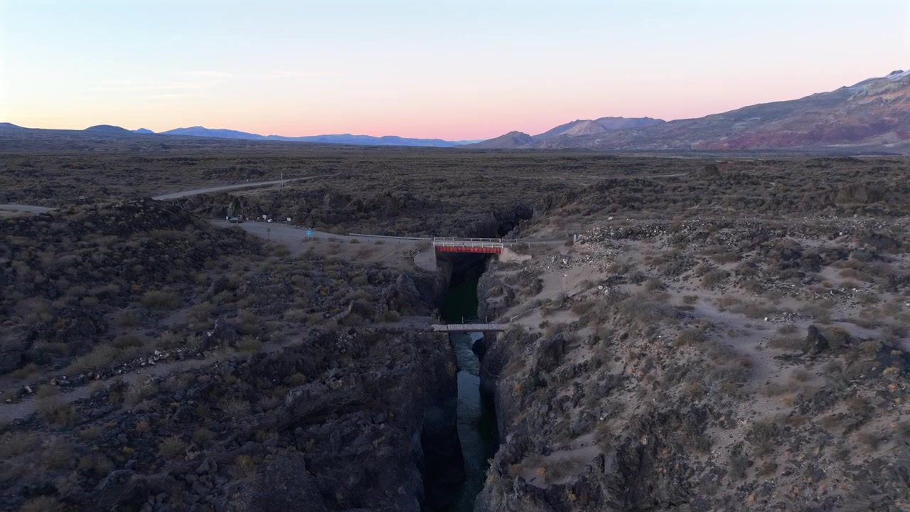 Volcanic canyon cuts through La Payunia Reserve in Mendoza, Argentina, revealing basalt formations and a bridge crossing the narrow gorge at sunrise, drone parallax pullback ascent shot.