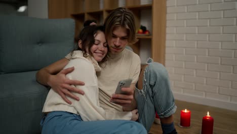 Romantisches-Paar-Sitzt-Zusammen-Auf-Dem-Boden-Und-Schaut-Auf-Den-Smartphone-Bildschirm