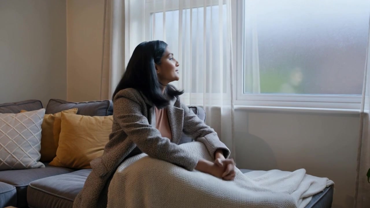Donna asiatica in cappotto che entra, cammina verso una chaise longue, si siede e drappeggia una coperta, guardando fuori dalla finestra