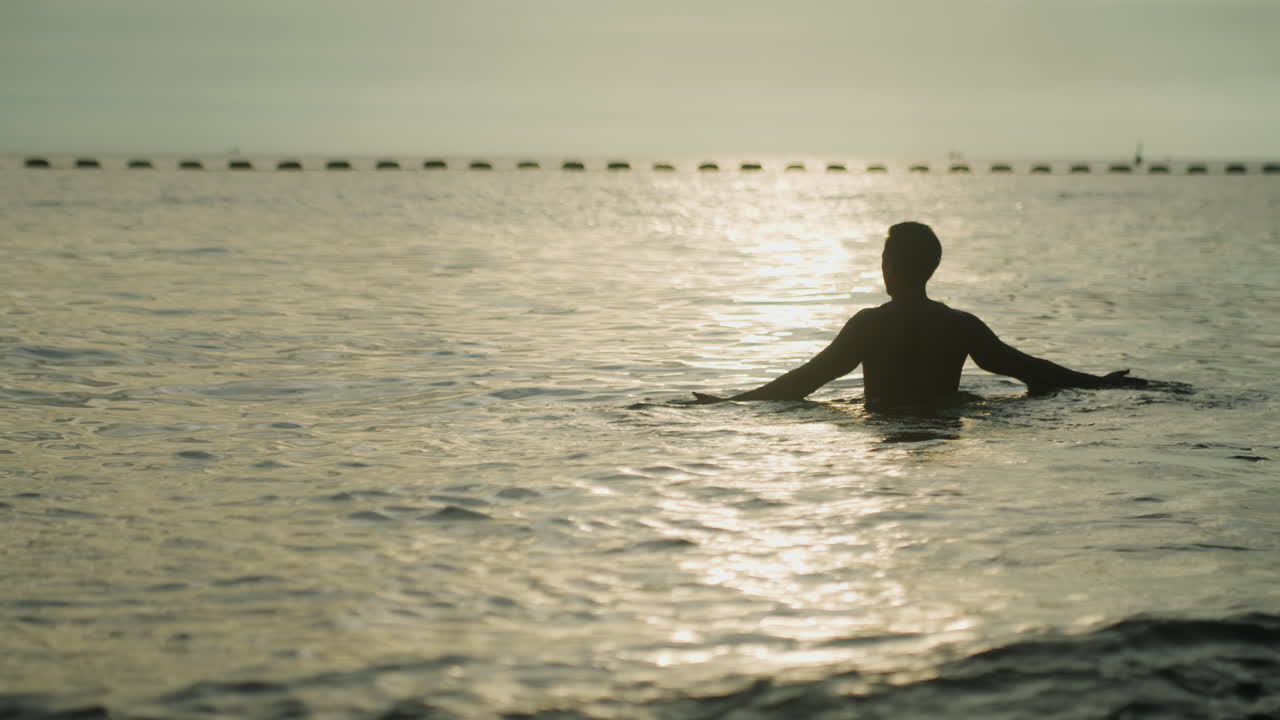 Silhuet af mand, der vader i havet ved solopgang, eftertænksom tidlig morgensvømmer, der vender mod horisonten med baggrundsbelyst profil, gyldent skær på vandet, blide krusninger, meditativ stilhed, eftertænksom stemning.