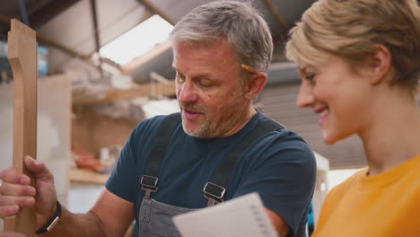 female apprentice making notes learning from mature male carpenter in furniture workshop