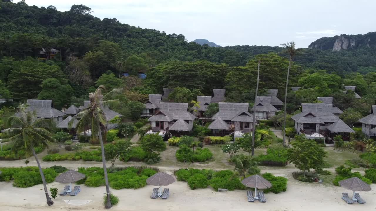Aerial view of SAii Phi Phi Island Village in Thailand, showcasing charming beachfront bungalows nestled amidst lush greenery and towering hills, with beach chairs and umbrellas.