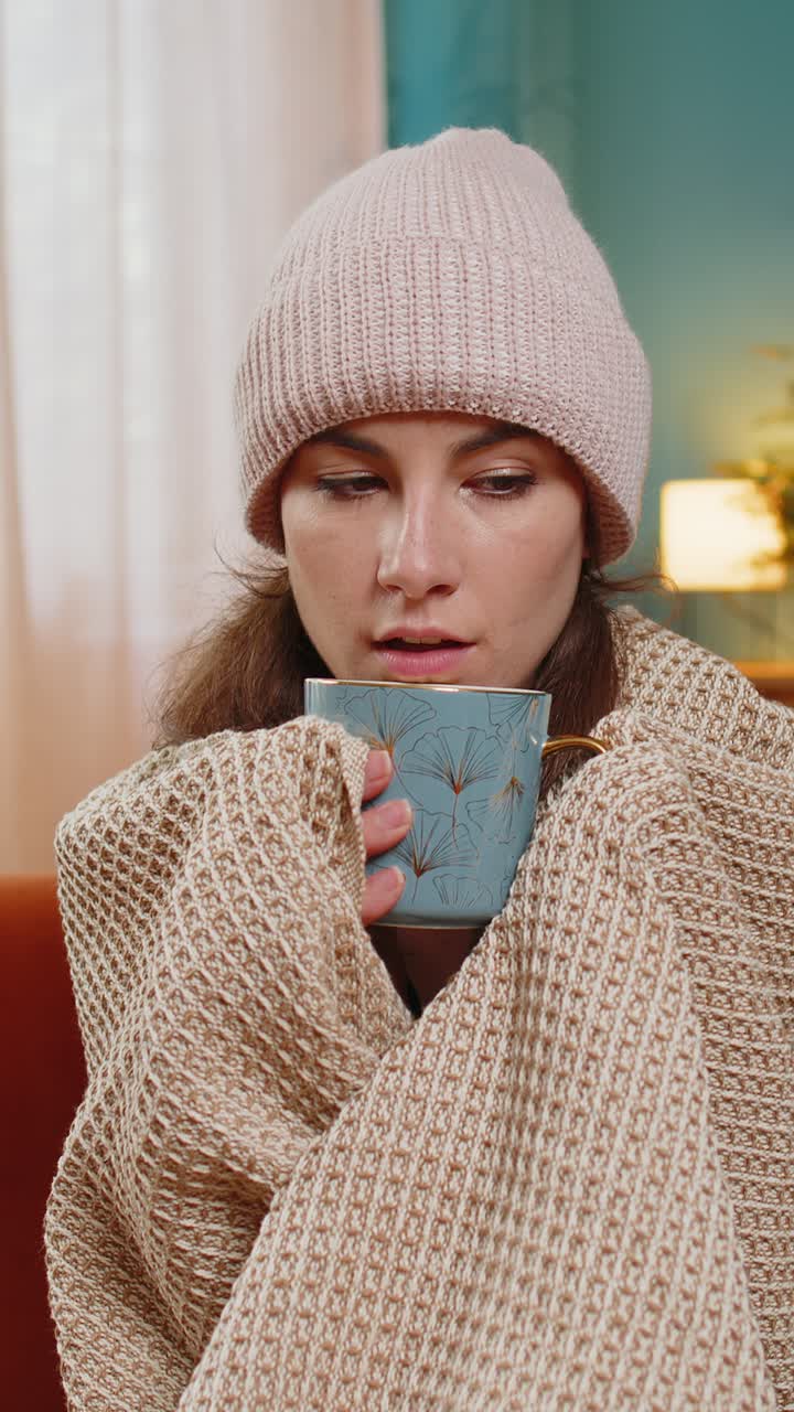 Young unhealthy sick ill woman covered by warm blanket drinks coffee freezes from cold flu at home