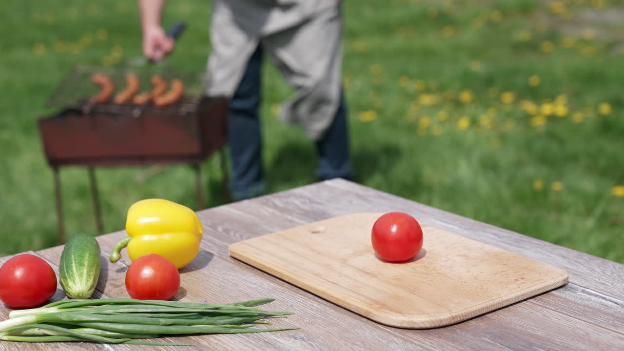 Fresh vegetables on wooden table outdoors. Food for picnic on the blur background of nature. Man frying sausages on a brazier.