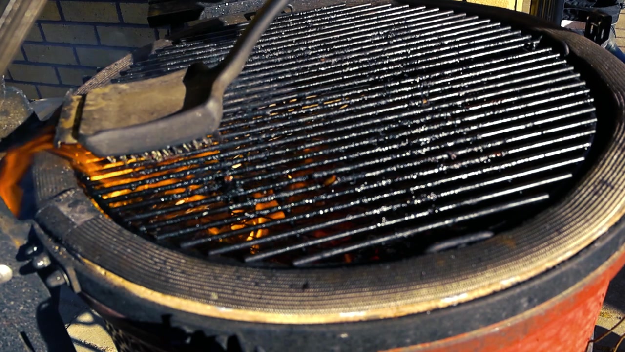 En person bruker en trebørste for å rengjøre den skitne metallristen på en varm kamado-grill, med flammer og røyk som stiger fra det brennende kullet under på en solrik sommerdag.