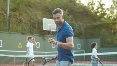 Schöner-Mann-Trainiert-Und-Schlägt-Ball-Mit-Schläger,-Während-Seine-Familie-Im-Hintergrund-Tennis-Spielt-1